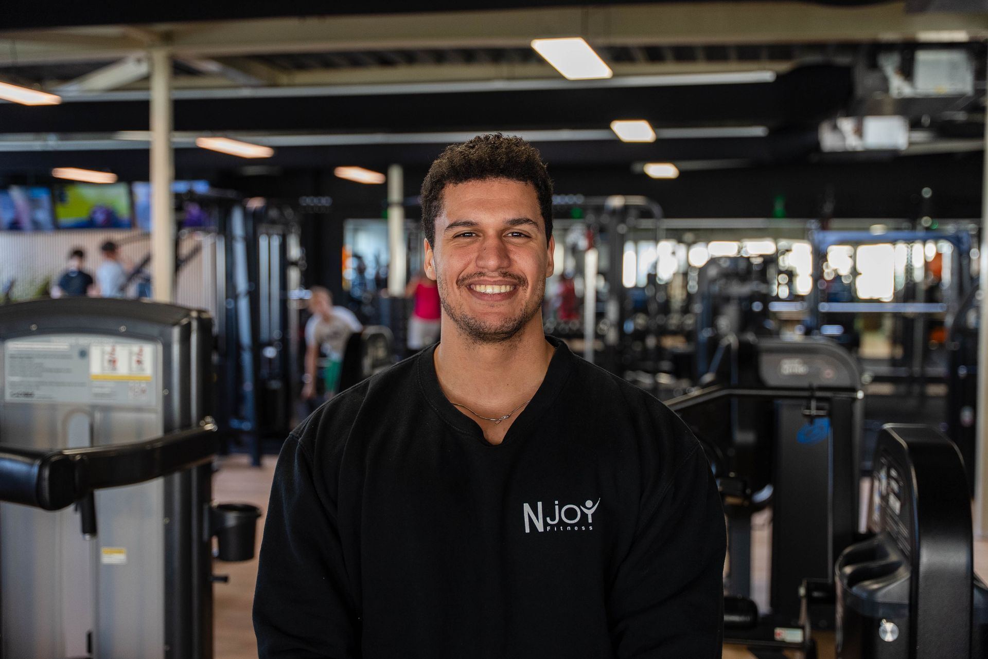 Een man in een zwart shirt lacht in een sportschool, omringd door fitnessapparatuur.