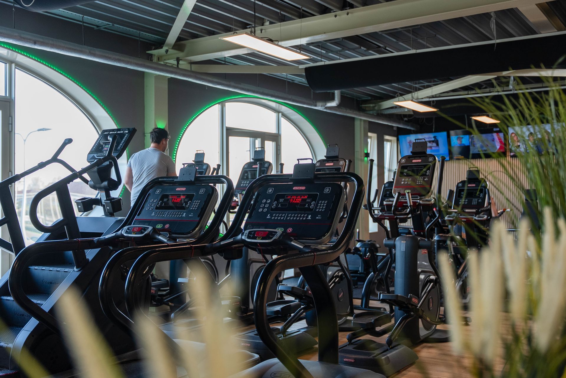 Interieur van een sportschool met crosstrainers, een persoon en grote boogvormige ramen.