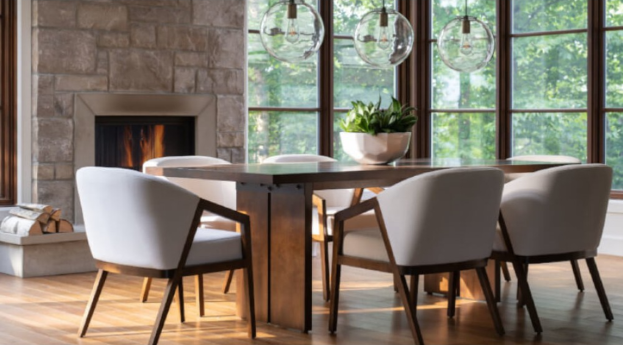 Dining room with table, chairs, fireplace, and large windows with a view of greenery.