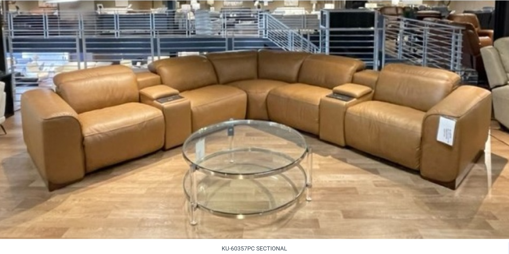 A curved, tan leather sectional sofa with cup holders, and a glass coffee table in a showroom.