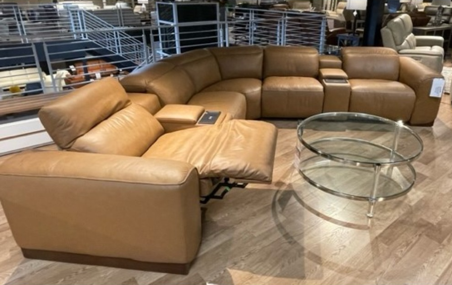 A curved, tan leather sectional sofa with cup holders, and a glass coffee table in a showroom.