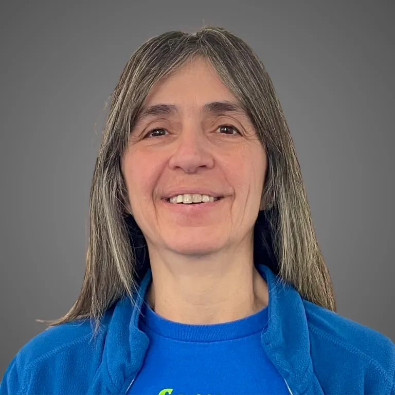 Woman with long graying hair smiles, wearing a blue fleece jacket over a blue top with a green design, in front of a gray backdrop.
