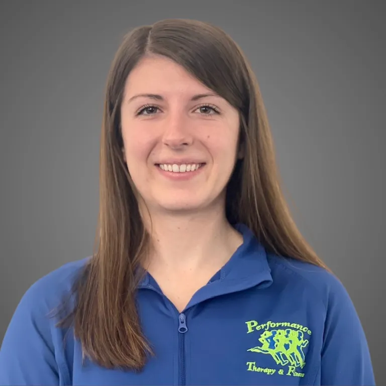 Woman in blue zip-up shirt, smiling. Performance Therapy & Pain logo on the shirt. Gray background.