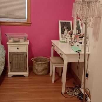 A bedroom with pink walls , a white desk , a lamp and a nightstand.