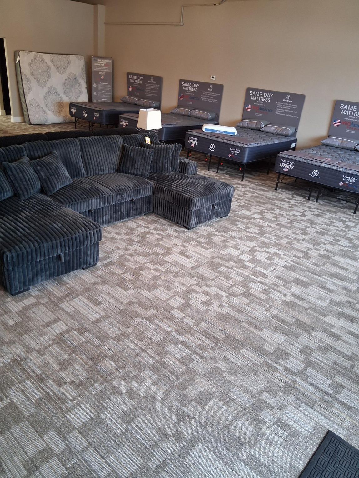 A showroom displaying dark gray sectional sofa, ottomans, and mattress sets on a patterned carpet.