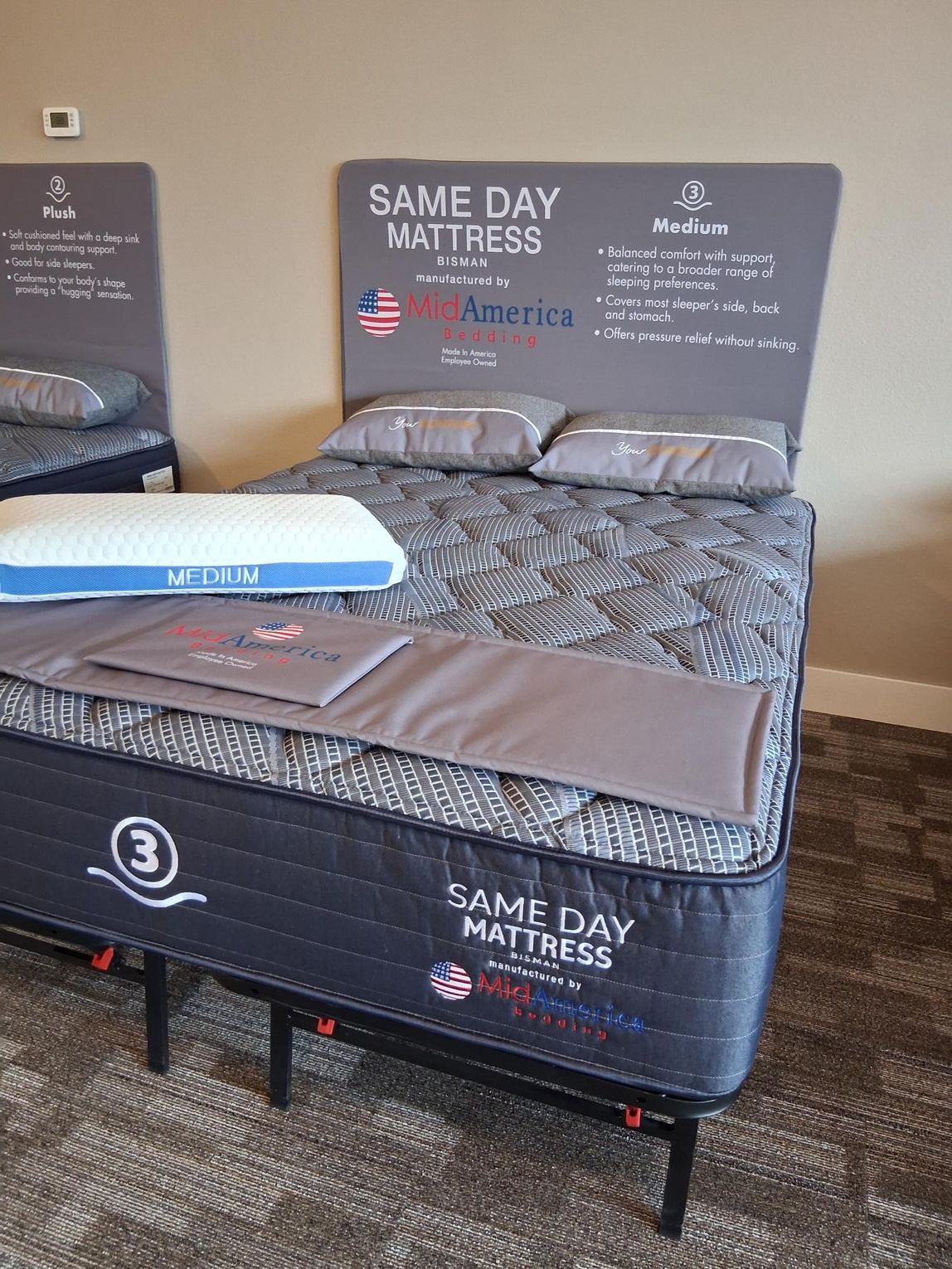 A display bed featuring a mattress with a gray and patterned top, pillows, and a pillow. Signage indicates 