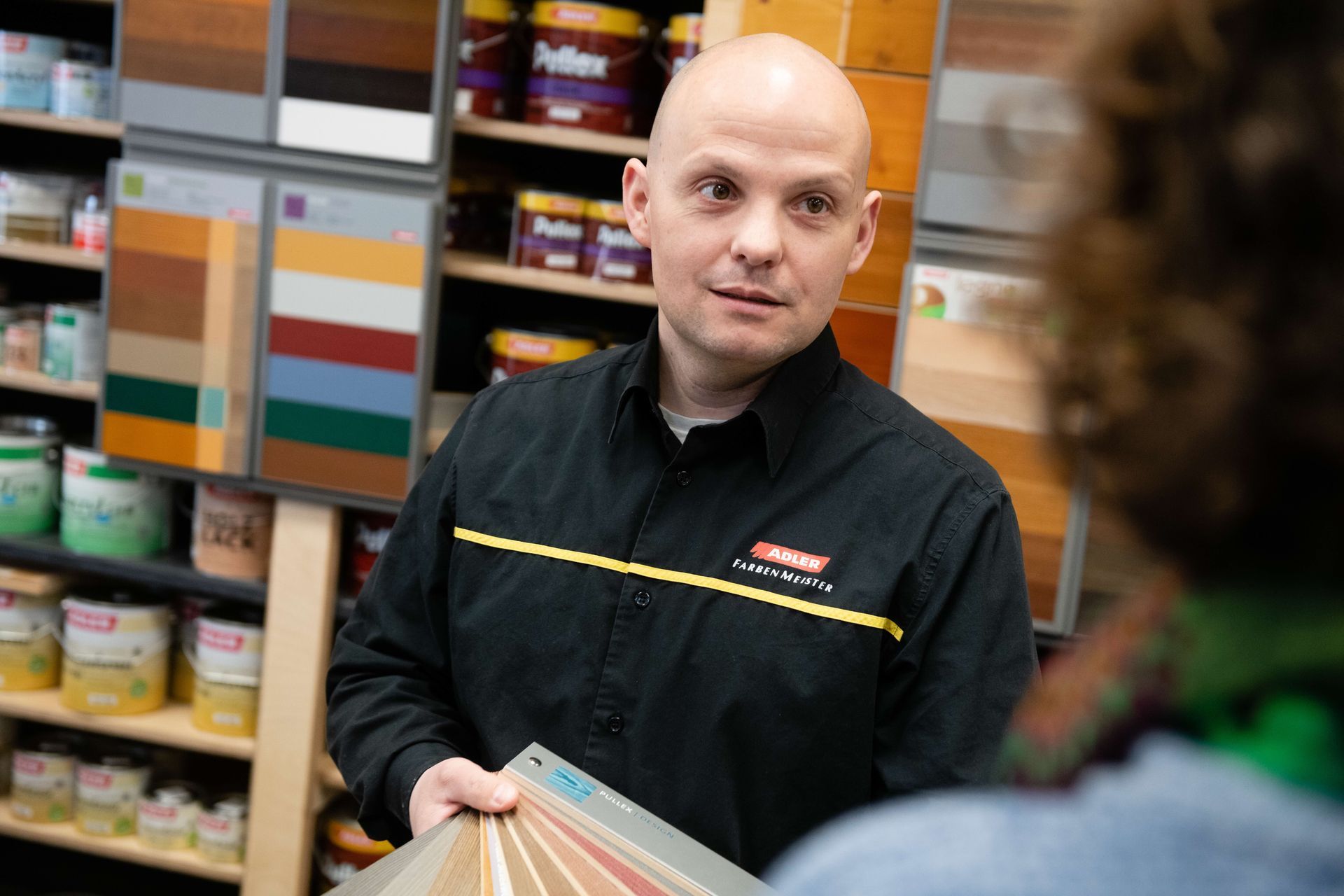 Ein Mann in schwarzem Arbeitshemd spricht in einem Farbengeschäft; hinter ihm sind Farbmuster und Regale zu sehen.