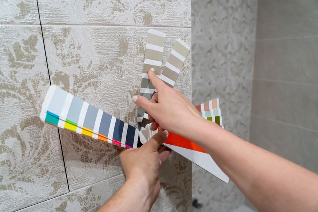 Hände halten einen Farbfächer an eine geflieste Wand und zeigen Farbmuster.