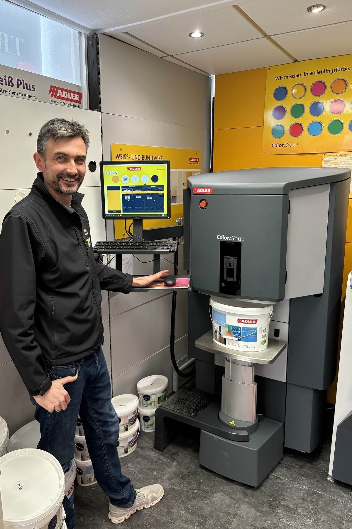 Ein Mann steht neben einer grauen Industriemaschine in einer Werkstatt; es handelt sich um eine Farbmischmaschine