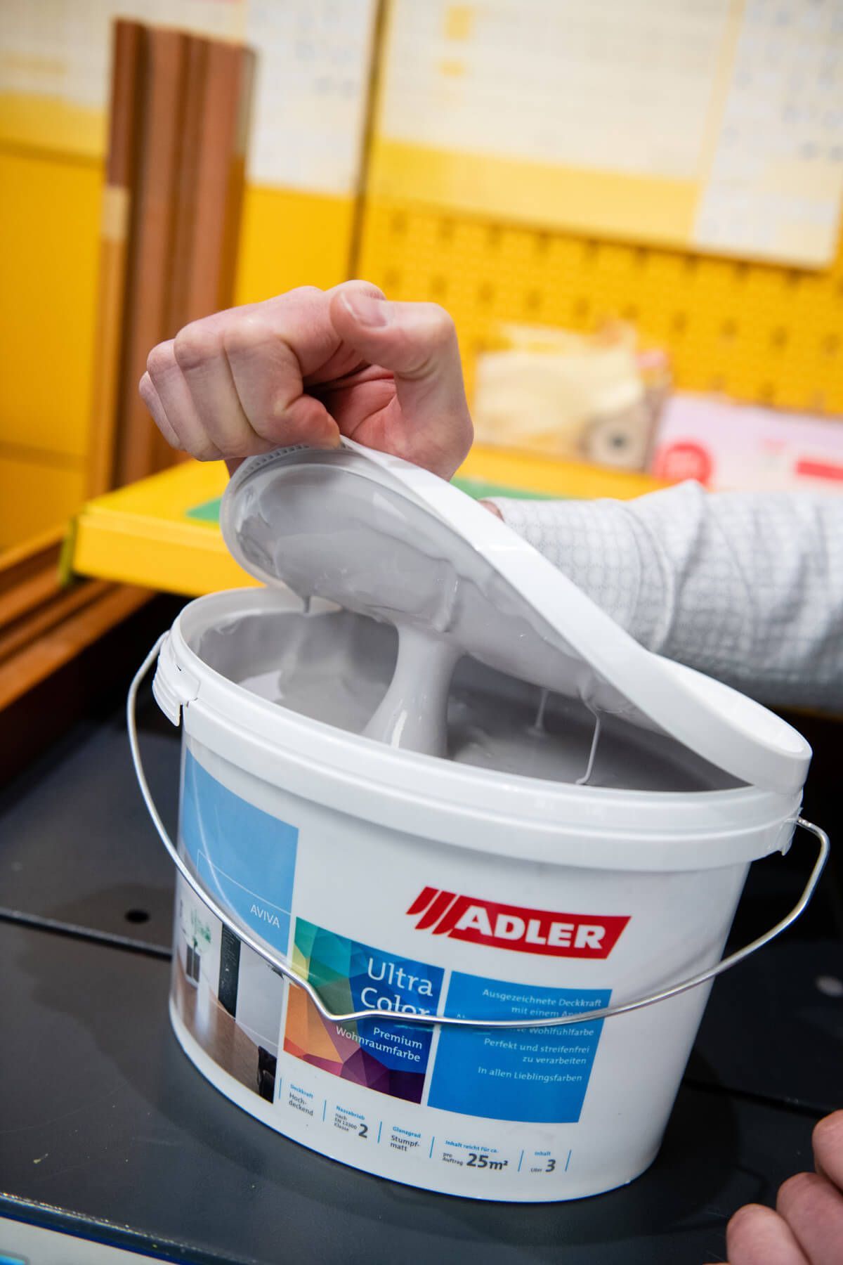Eine Hand hält den Deckel eines weißen Adler-Farbeimers in einer werkstattähnlichen Umgebung offen.