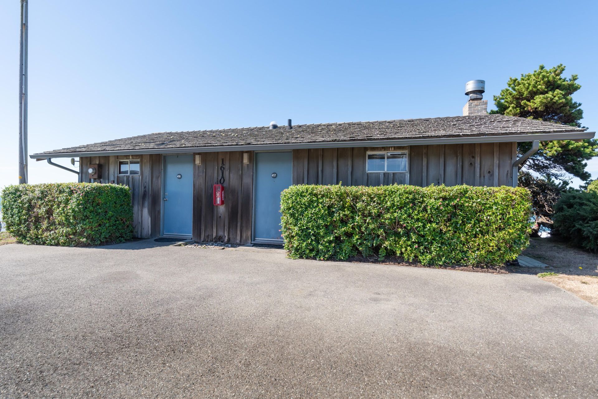 Wooden public restroom with blue doors and weathered roof, surrounded by green hedges, under a blue sky.