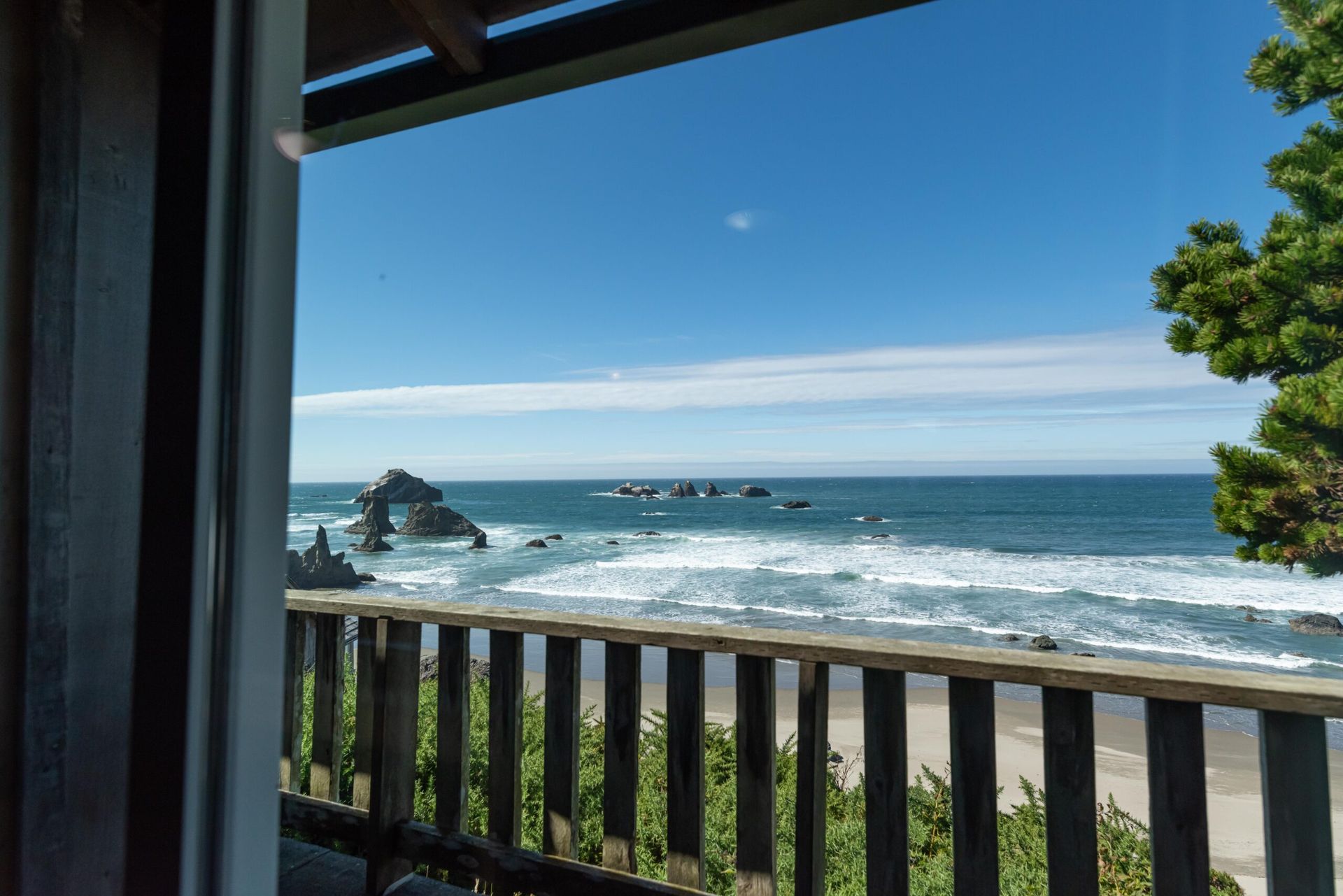 Ocean view from a wooden deck, blue sky and water, waves crashing on beach, rocks in the sea, green tree.