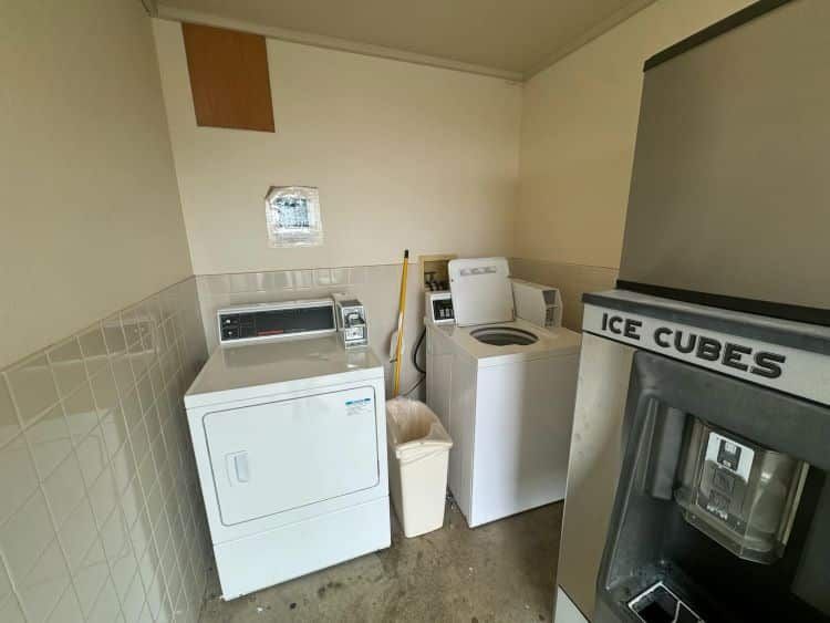 Laundry room with dryer, washing machine, ice machine, and trash can. Beige walls and tiled wainscoting.