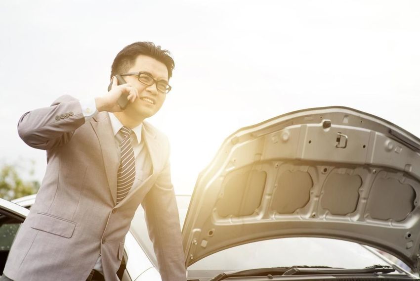 A man in a suit is talking on a cell phone next to a broken down car.