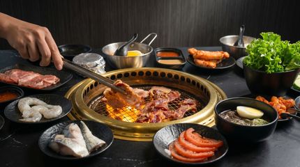 Hands cooking meat and seafood on a tabletop Korean barbecue grill with side dishes and lettuce