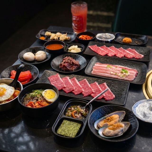 Assorted raw and cooked meats with side dishes on black trays, plus a red drink on a dark table.