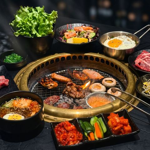 Korean barbecue table with sizzling grill, meats, side dishes, lettuce, and bowls of rice and soup