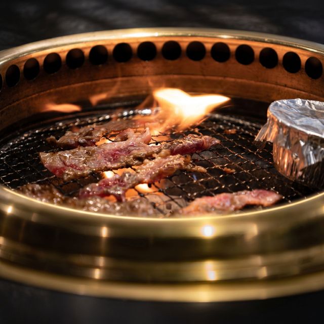 Beef sizzling on a tabletop grill with a small flame and metal foil cup nearby
