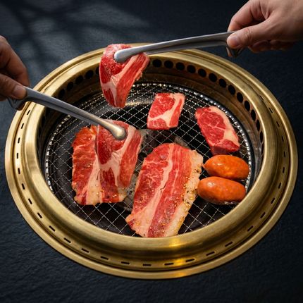 Hands placing marbled beef and sausages on a round tabletop grill with a brass rim