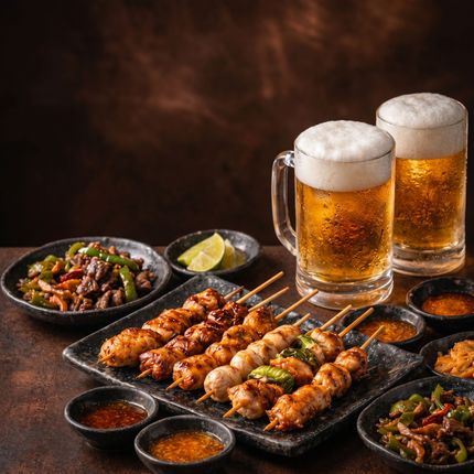 Grilled kebabs and side dishes with two frothy beers on a dark table