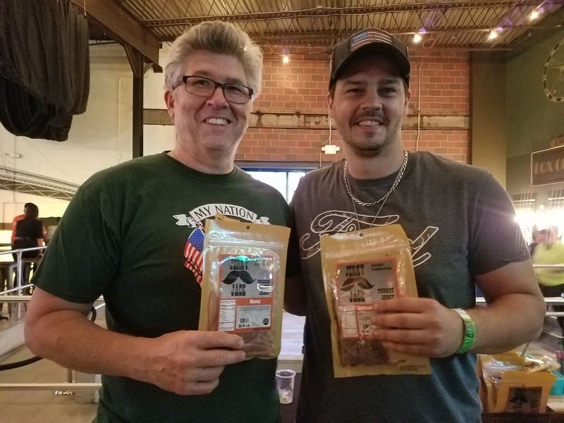 Two men are standing next to each other holding bags of jerky.