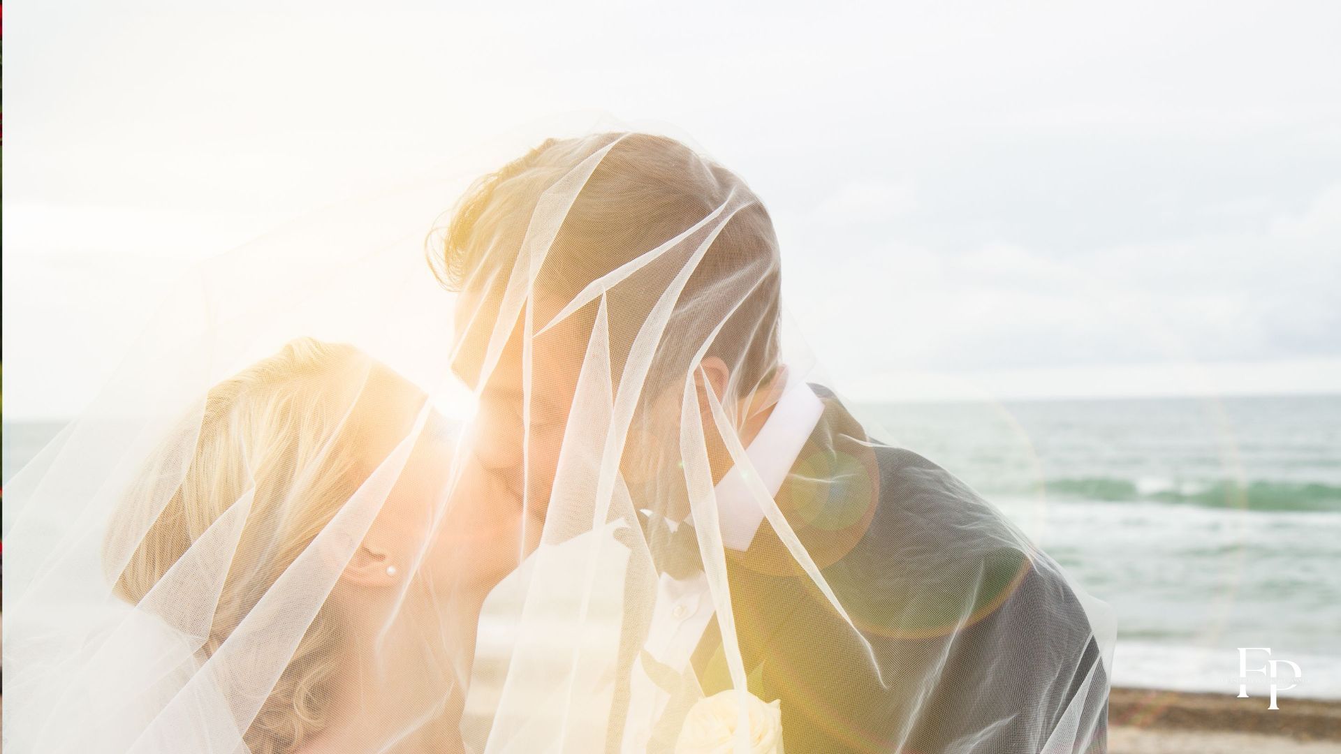 The bride's radiant smile illuminates her face as she leans into her partner, her veil gently fluttering in the evening breeze, while groom, dapper in his suit, cradles her face tenderly.
