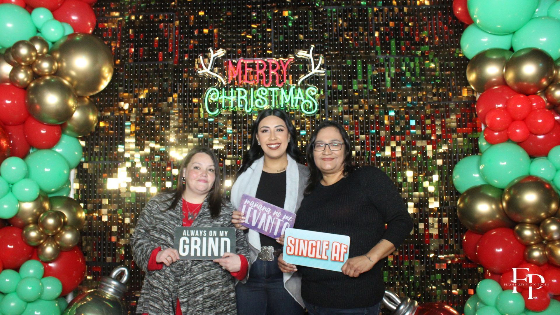 Three smiling women posing with a Christmas sign inside the Oval Mirror Photo Booth.