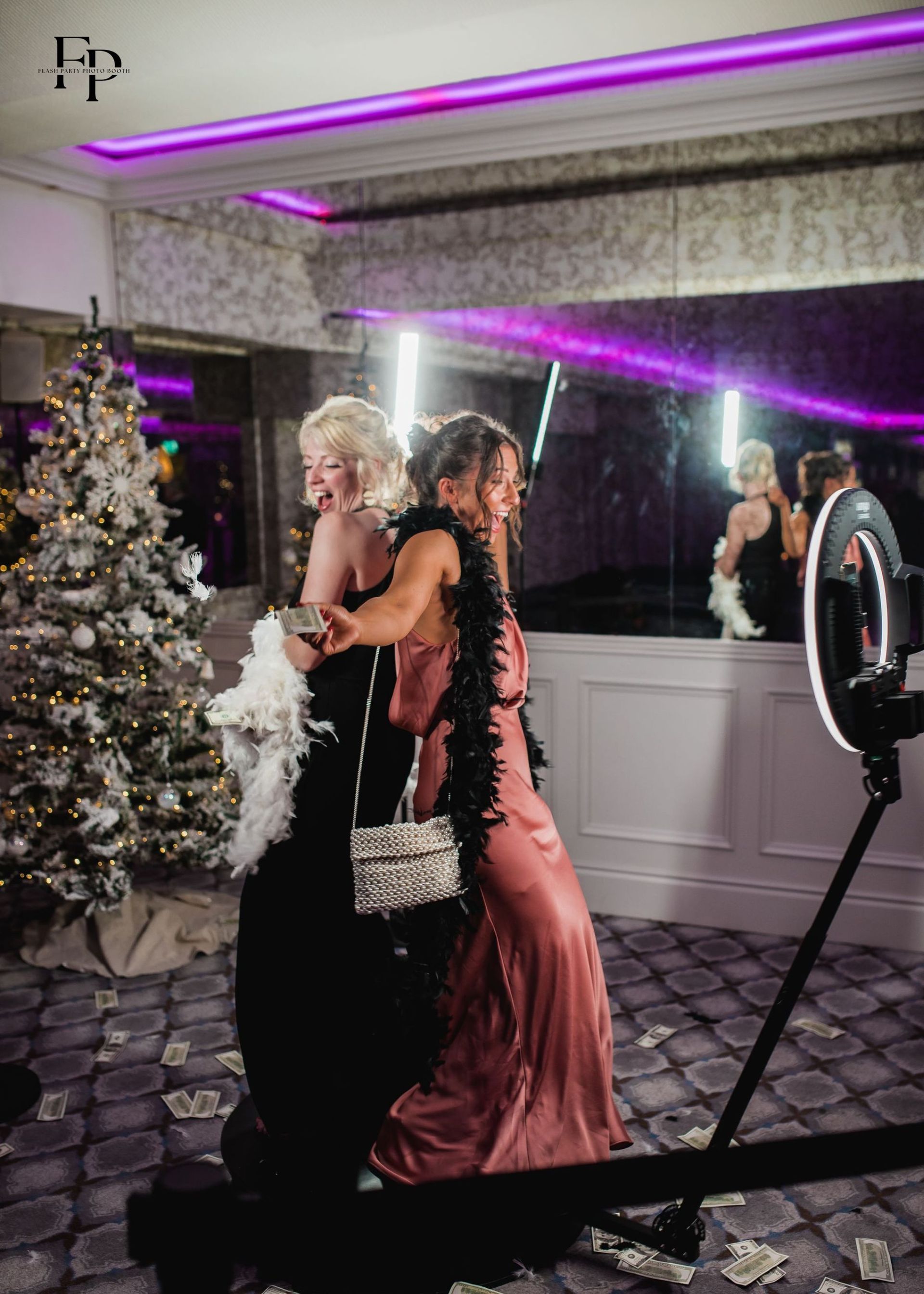 Two women in fancy gowns posing by a festive Christmas tree in front of 360 Photo Booth.