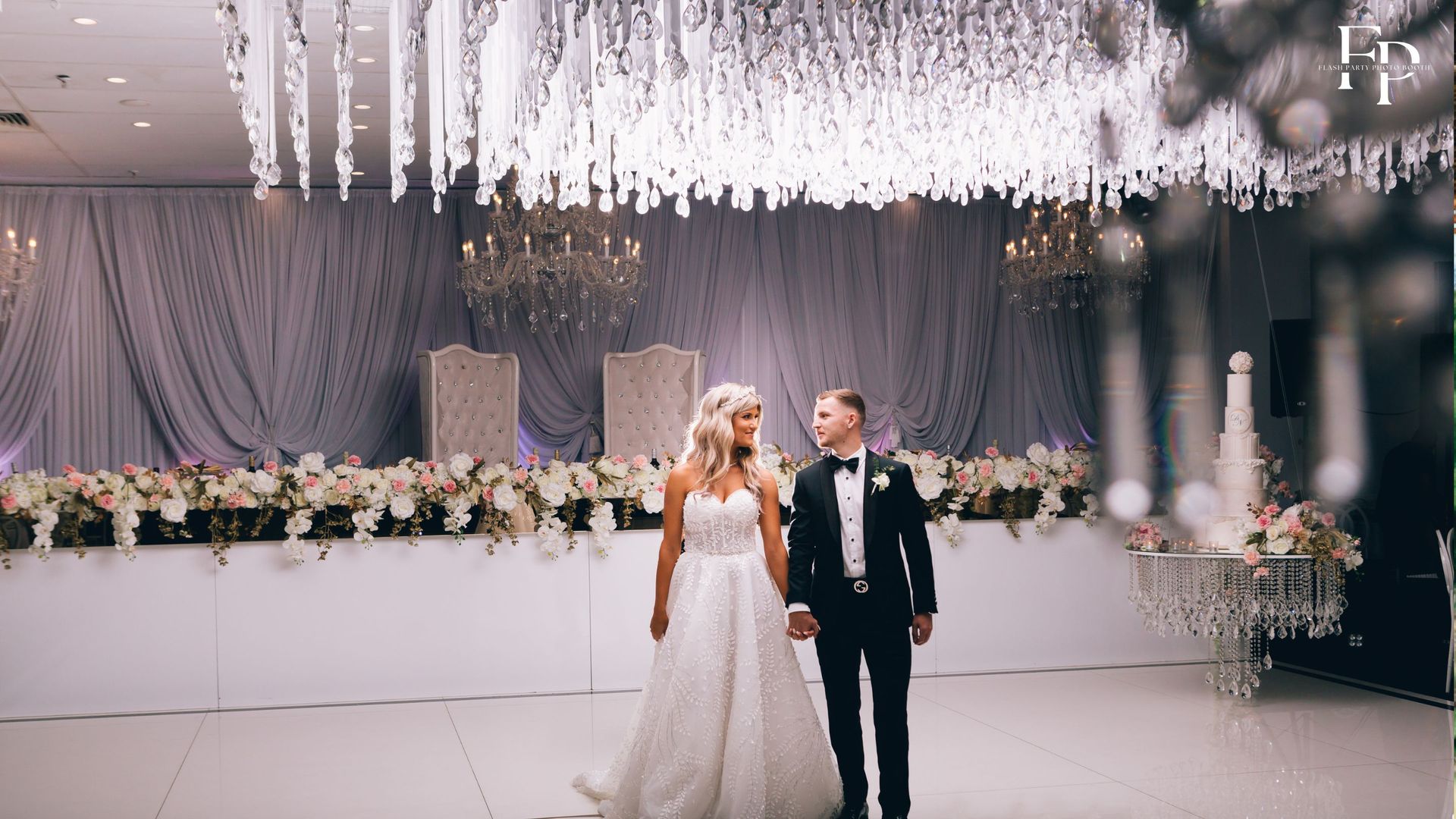 Surrounded by opulent elegance, the bride and groom stand at the center of the grand reception hall in DFW, their love radiating like a beacon of light amidst the festive atmosphere.