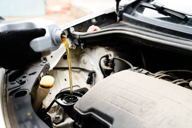 Person pouring motor oil into a car's engine from a gray container, engine bay visible.