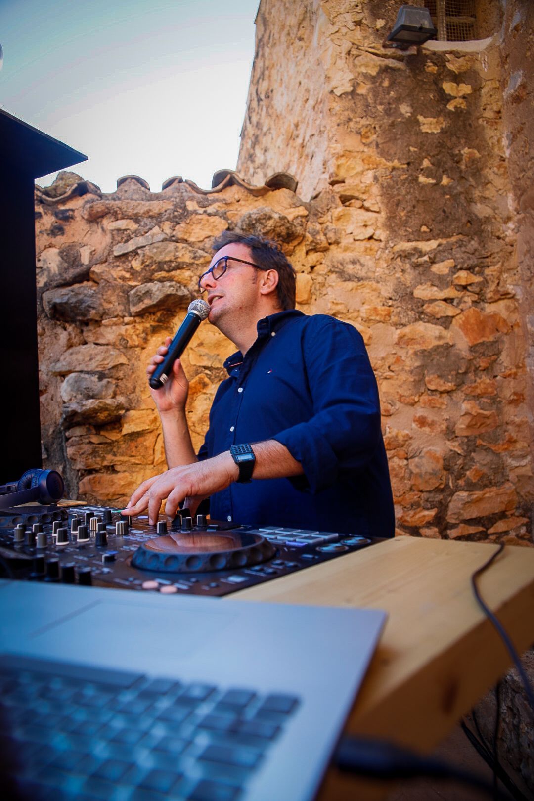DJ con camisa azul y micrófono, mezclando en un evento al aire libre cerca de un muro de piedra.