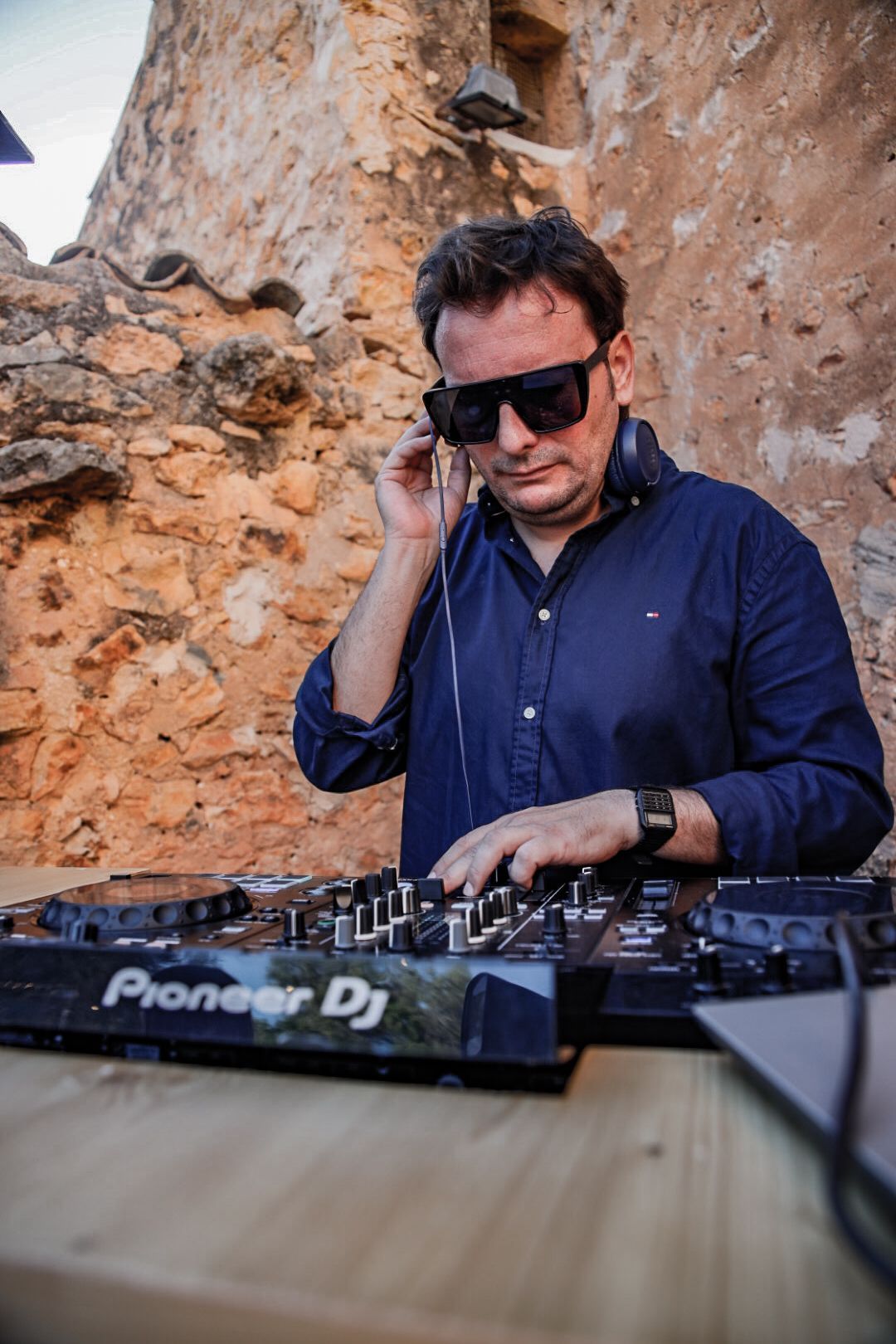 DJ con gafas de sol y auriculares, trabajando en un mezclador Pioneer DJ, al aire libre cerca de un muro de piedra.