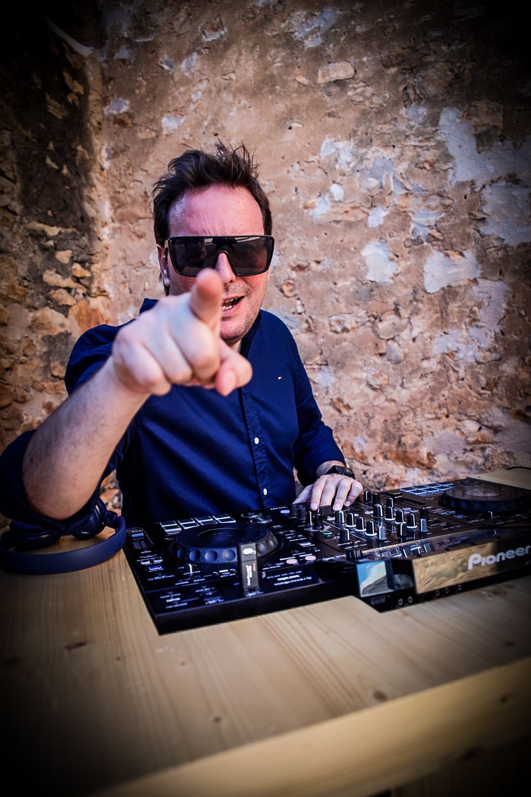 Un DJ con gafas de sol señala a la cámara. Está en una mesa de mezclas Pioneer en el exterior.