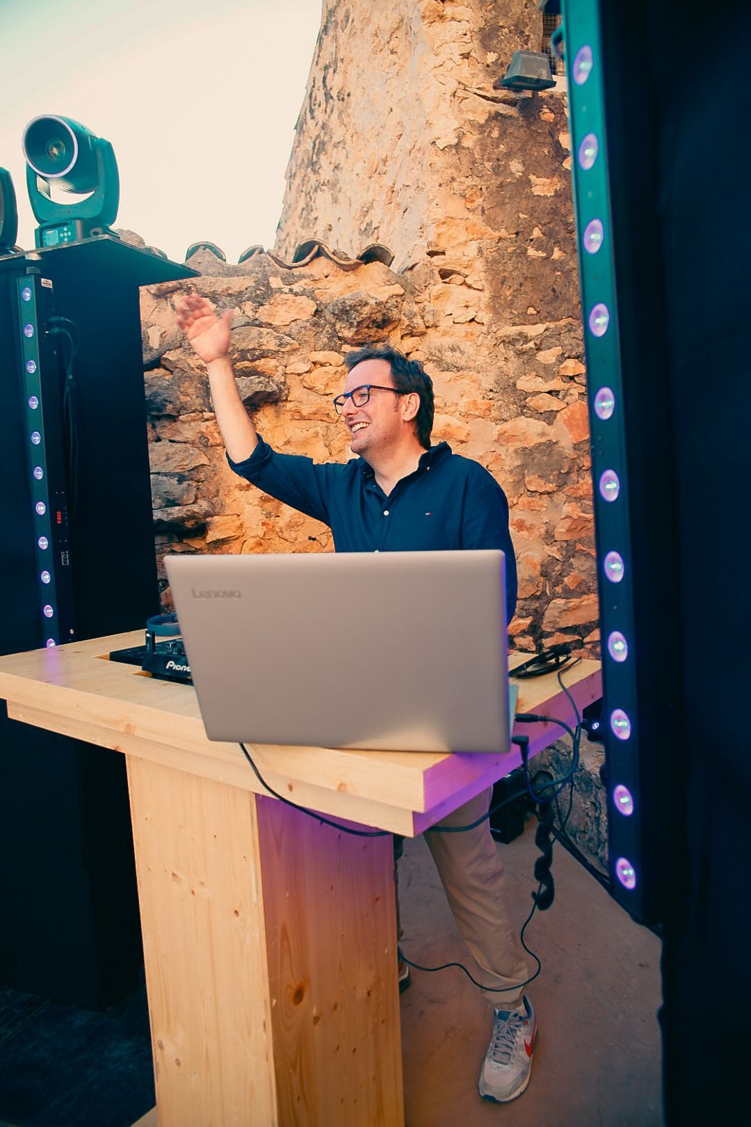 DJ en un evento al aire libre con su portátil sobre una mesa de madera. Contra un muro de piedra, con altavoces e iluminación