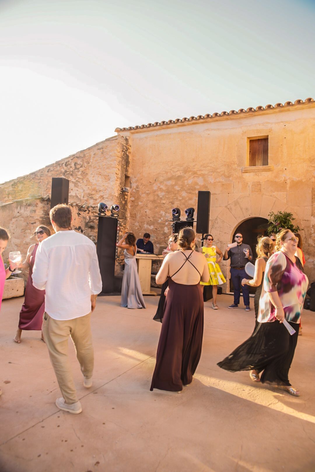 Gente bailando en un evento al aire libre cerca de un edificio de piedra. DJ al fondo, iluminado por el sol.