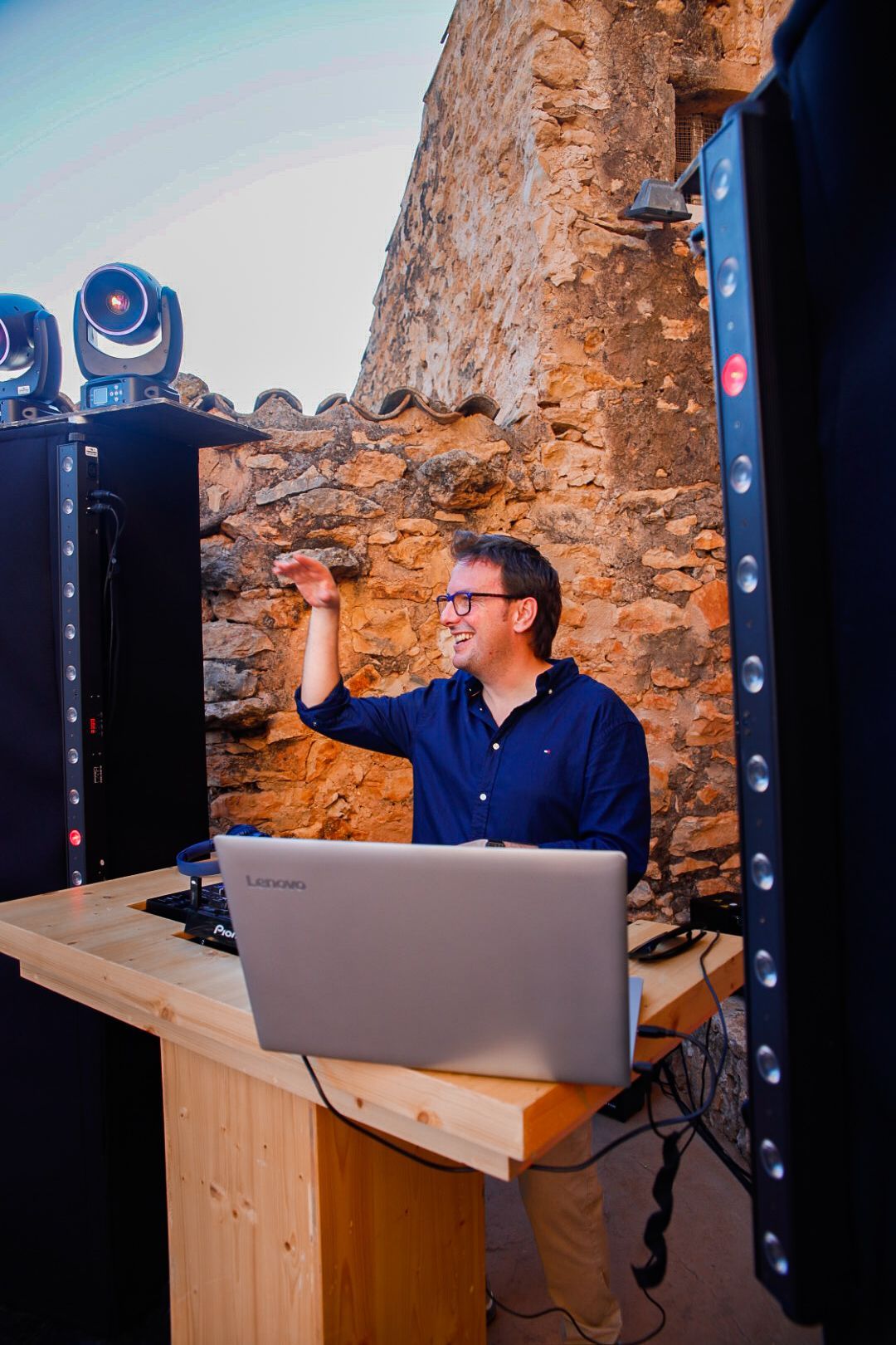 DJ sonríe, levanta el brazo, en una mesa de madera con computadora portátil, parlantes y luces, contra una pared de piedra.