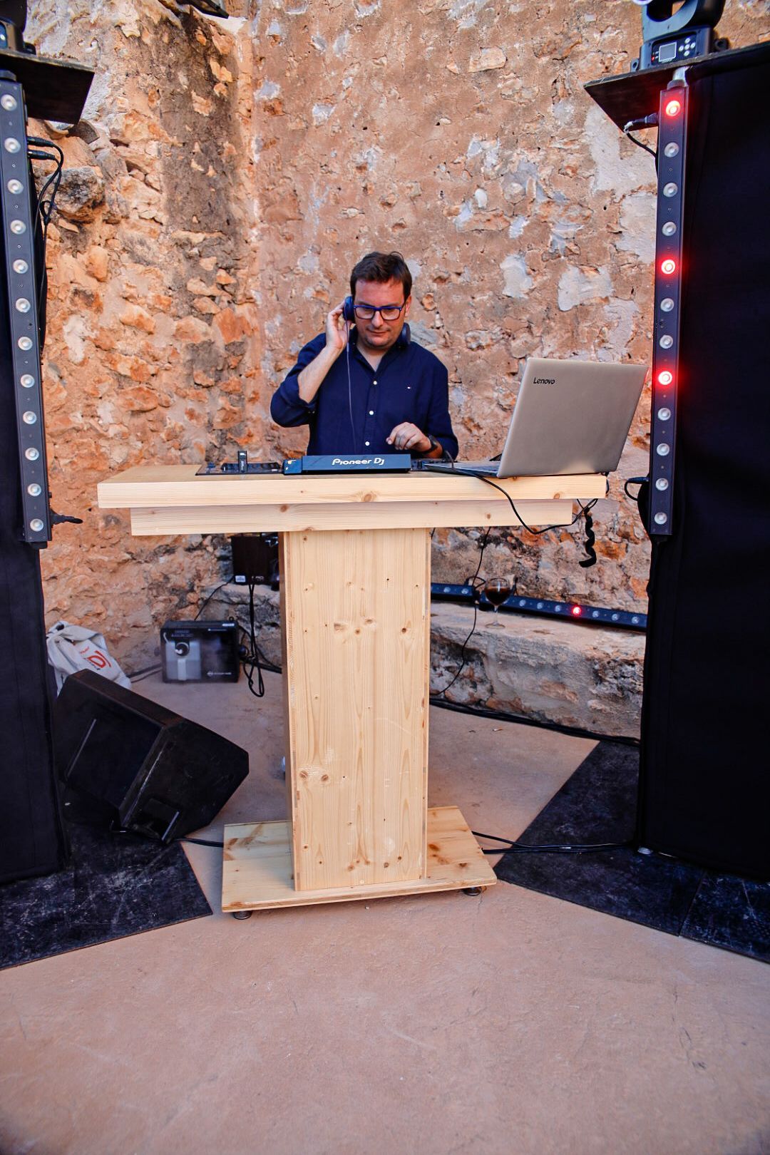 DJ con camisa azul y gafas en una mesa de madera frente a un muro de piedra.