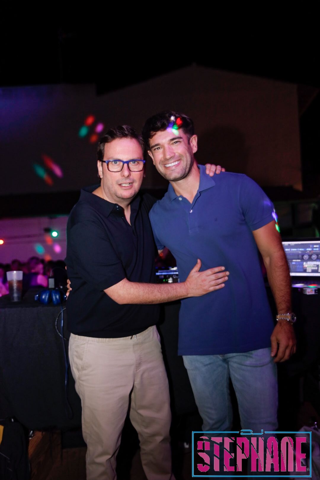 Dos hombres sonrientes, uno con gafas y pantalones caqui, el otro con camisa azul y vaqueros, posando en una fiesta.