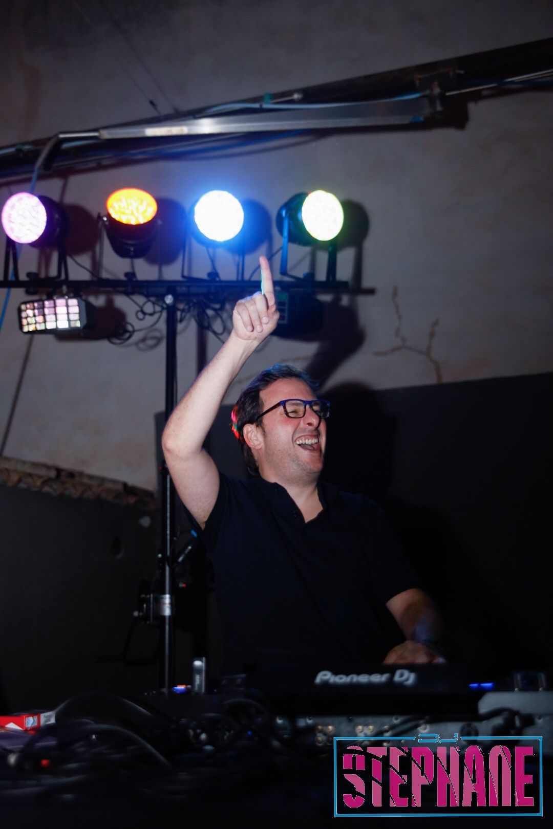 Hombre pinchando, señalando hacia arriba, sonriendo. Escenario con luces de colores. El logo dice 