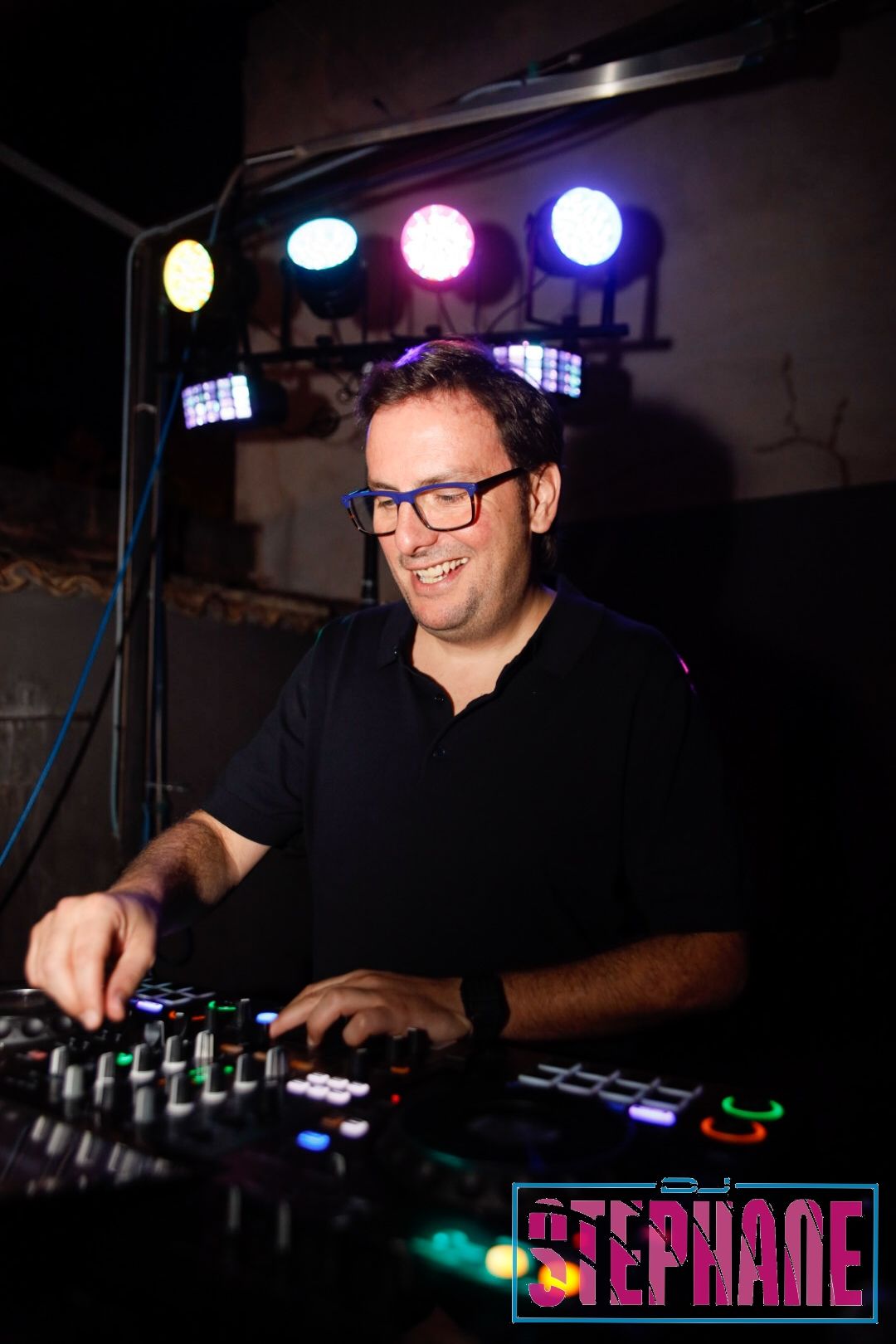 DJ Stéphane en un club, con gafas, sonriendo mientras mezcla música en una consola con luces de colores de fondo.