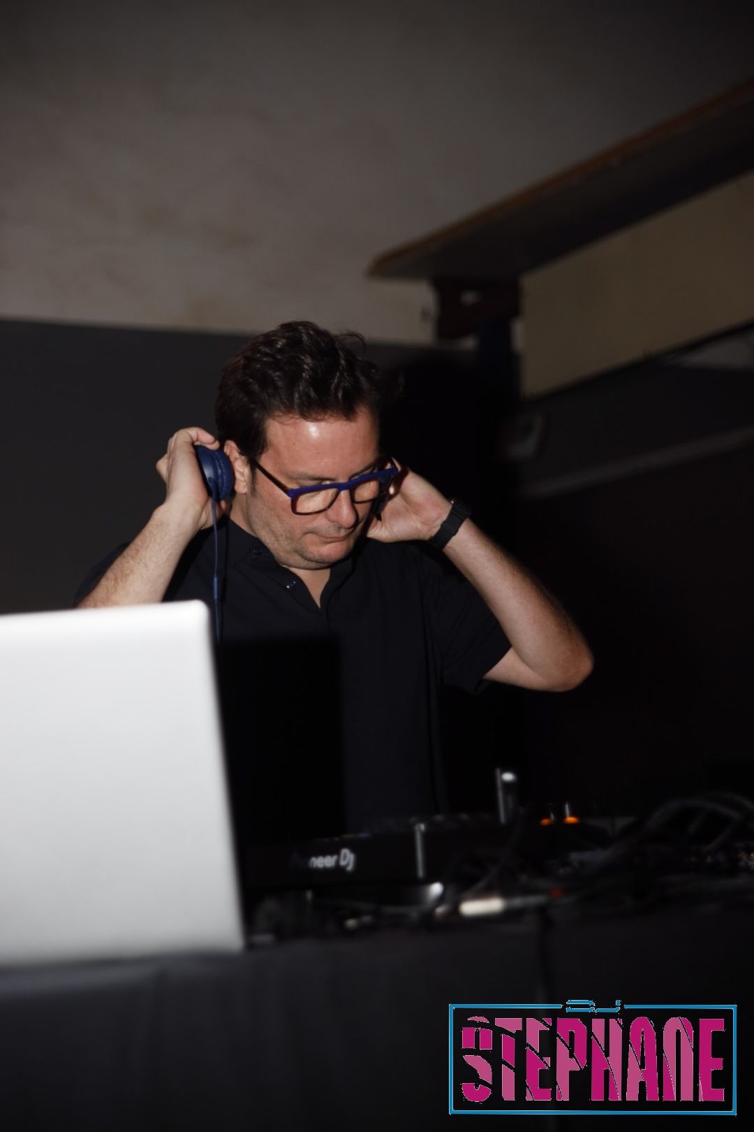 DJ Stephane ajustando los auriculares en una consola de mezclas con una computadora portátil en una habitación oscura.