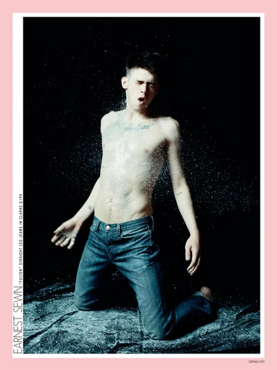 Shirtless person kneels, wet, mouth open. Dark background, light reflecting. Denim jeans.