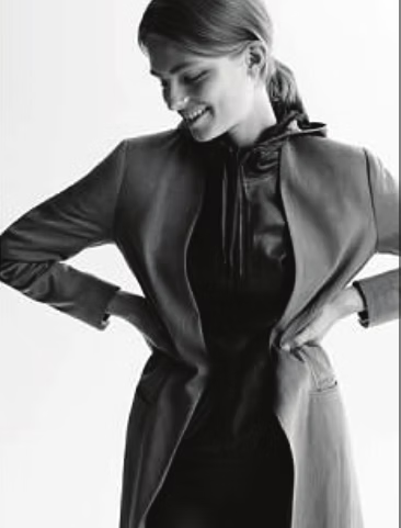 Woman smiling, wearing a dark coat and dress, hands on hips. Black and white photo.