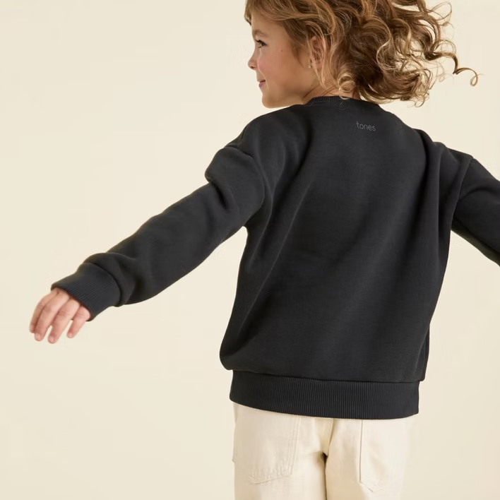 Child wearing a dark sweatshirt and tan pants with arms outstretched, against a light background.