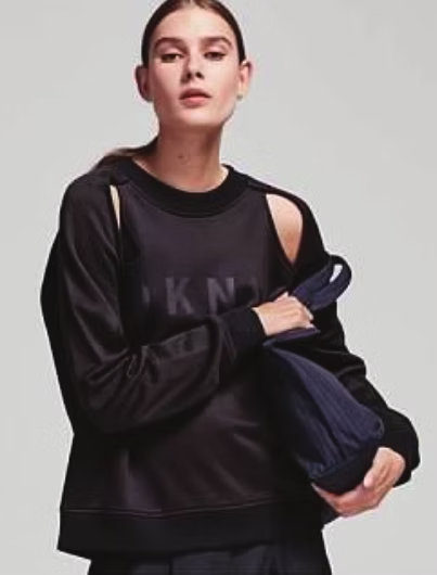 Woman in black sweatshirt with shoulder cutouts, holding a blue bag, against a white backdrop.