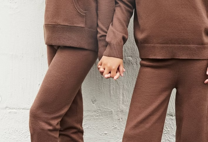 Two people in matching brown knitwear holding hands in front of a textured wall.