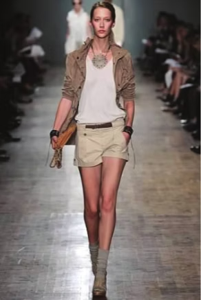 Model in tan outfit walks the runway, beige jacket, shorts, top, and booties, carrying a handbag.