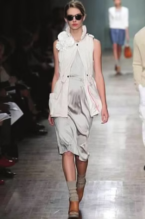 Model in light gray dress, white vest, sunglasses, on a runway.