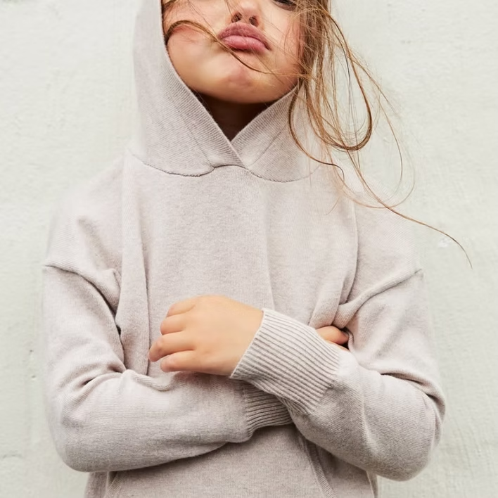 Girl in a light beige hooded sweater, arms crossed, face with pursed lips, against a white wall.