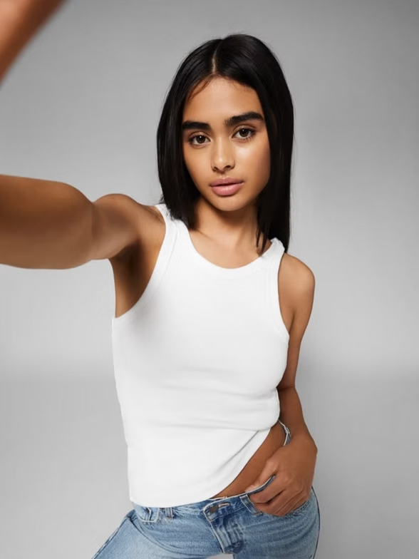 Woman in white tank top taking a selfie. Light background.